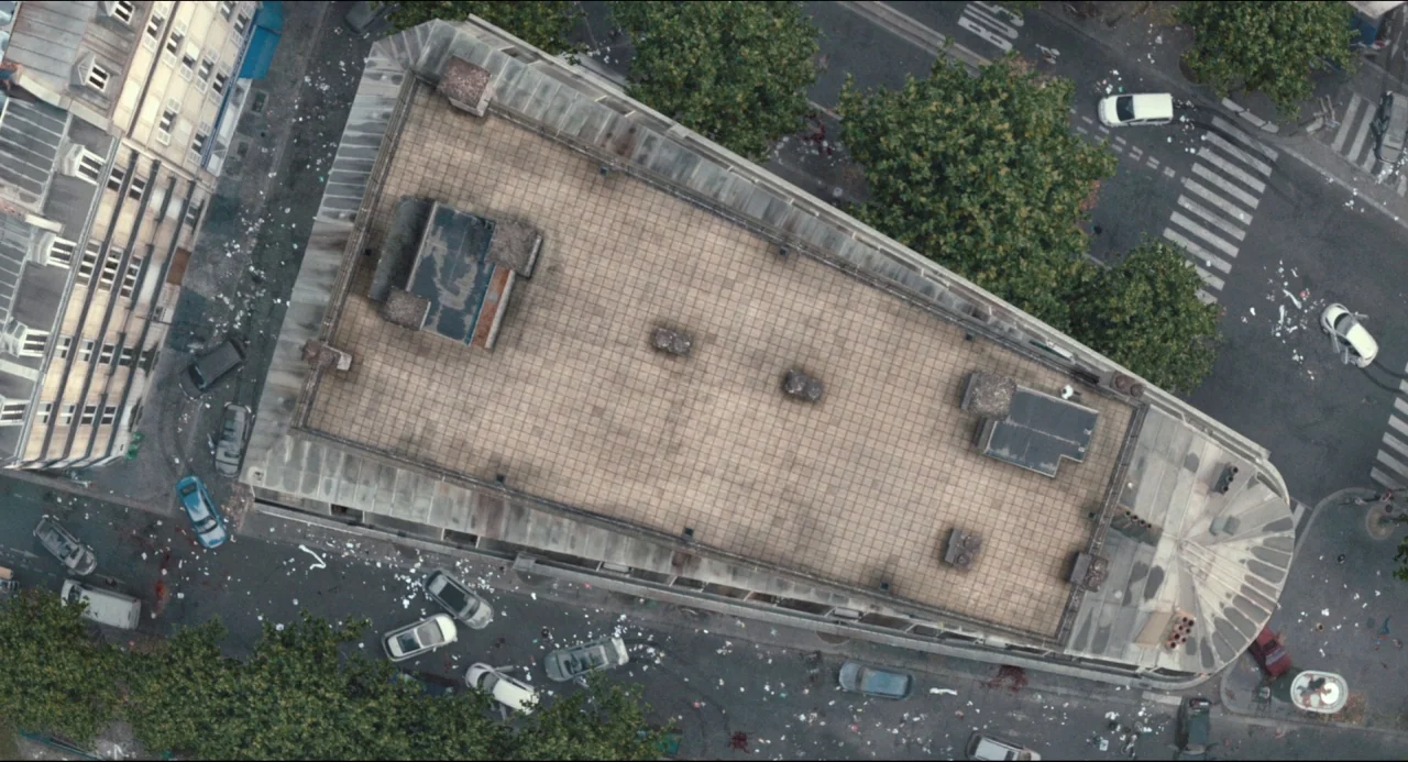 The Night Eats the World (2018) movie still: establishing — A high-angle top-down view shows an urban street corner with a large building rooftop.…