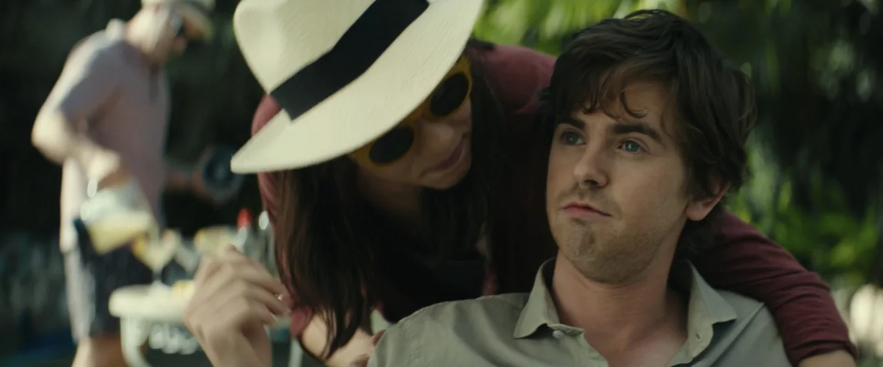 The Vault (2021) movie still: medium — An outdoor space featuring tropical foliage and a table with drinks in the background. A woman in a sun hat…