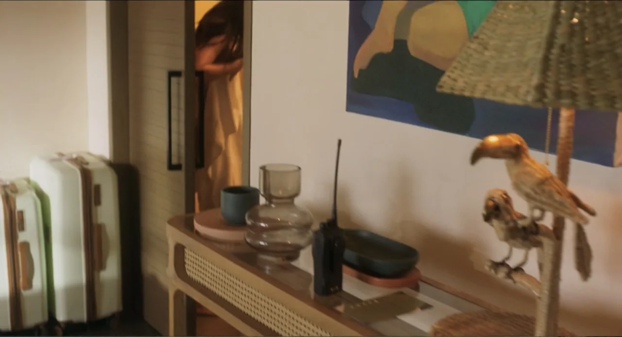 At Midnight (2023) movie still: medium — A hotel room features a rattan console table with decorative bird statues and glass vases. A woman wrapped…