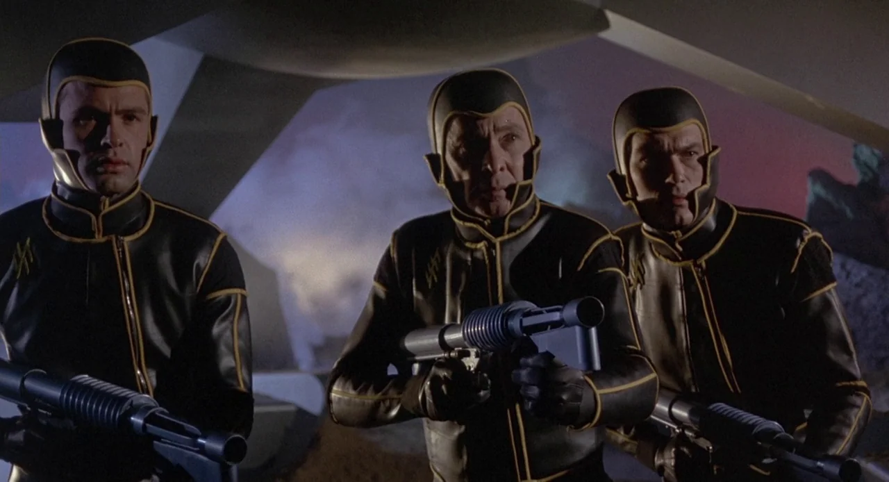 Planet of the Vampires (1965) movie still: medium — Three men stand on a dark, rocky terrain wearing matching black tactical uniforms. Each man…