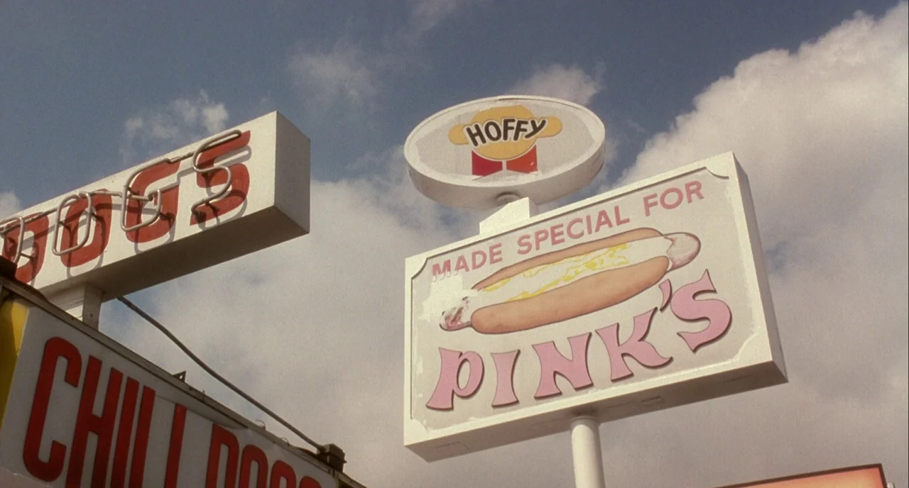 Mulholland Drive (2001) movie still: establishing — Several large commercial advertising signs for a hot dog restaurant stand against the sky. The…