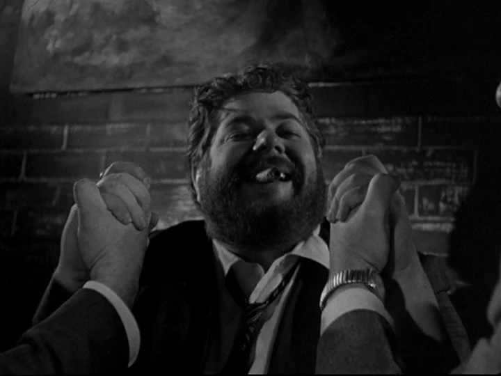 Blast Of Silence (1961) movie still: closeup — A dark indoor space features a brick wall background illuminated by sharp light. A bearded man in a…