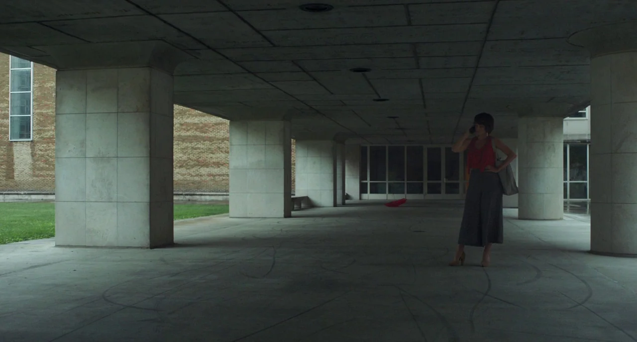 Columbus (2017) movie still: wide — A modernist covered outdoor walkway features massive concrete pillars and a tiled ceiling. A woman in a red top…
