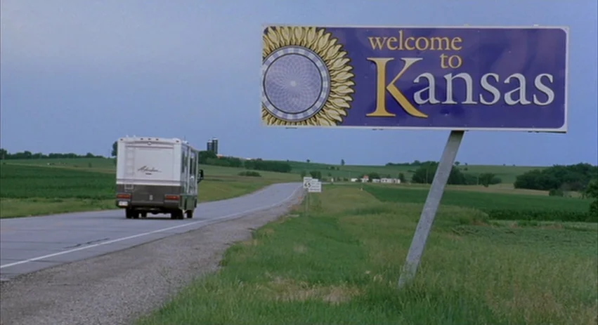 About Schmidt movie still: wide — A rural asphalt highway winds through green rolling fields under an overcast sky. A large welcome to Kansas sign…