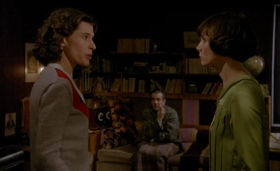 Mélo (1986) movie still: medium — Two women stand in a dimly lit room with bookshelves and a seated man. The space contains dark walls and a…