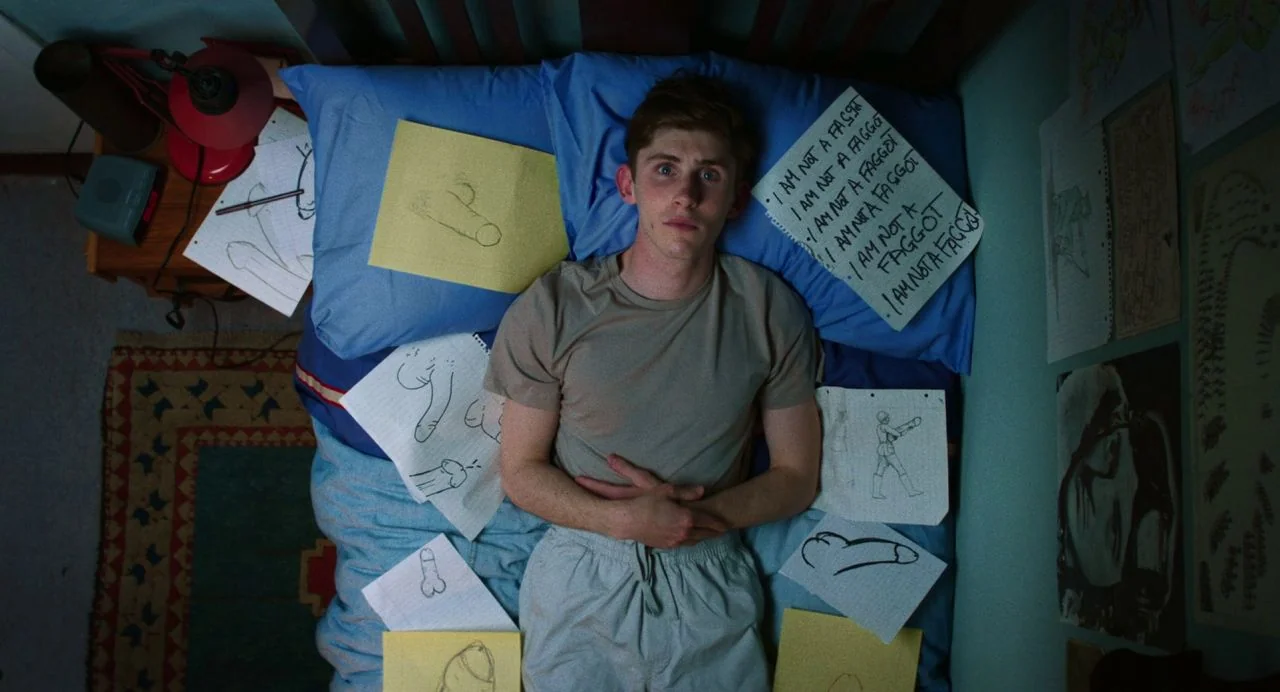 Dating Amber movie still: fullbody — A teenage boy lies flat on his back on a bed covered in drawings. The bedding is blue and several papers with…