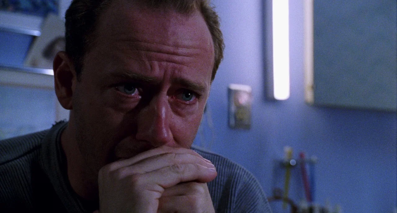 Candyman (1992) movie still: closeup — A dimly lit bathroom features blue-toned walls and a vertical fluorescent light fixture. A middle-aged man…