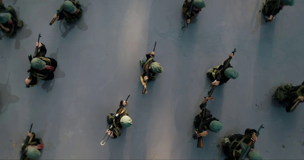 First They Killed My Father (2017) movie still: wide — A group of armed individuals marches across a flat, grey asphalt surface. They wear tactical…