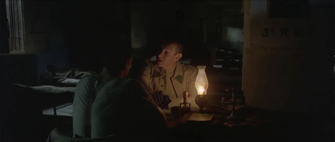 Legionario (2025) movie still: medium — A dimly lit room serves as a military office with simple furnishings and dark walls. One man in uniform…