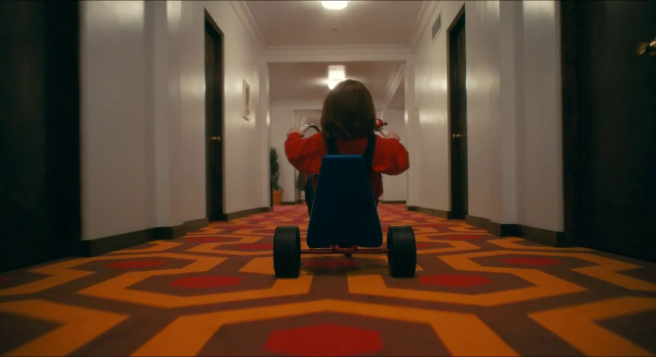 Doctor Sleep movie still: fullbody — A long symmetrical hotel hallway features geometric orange, red, and brown carpet patterns. A young boy rides a…