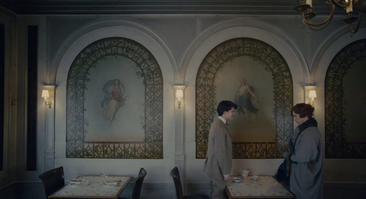 The Danish Girl (2015) movie still: wide — An interior restaurant room features arched murals of figures on the walls. A man and a woman stand by a…