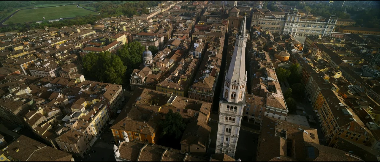 Ferrari (2023) movie still: establishing — An aerial perspective captures a dense European city center featuring a tall white bell tower. Rows of…