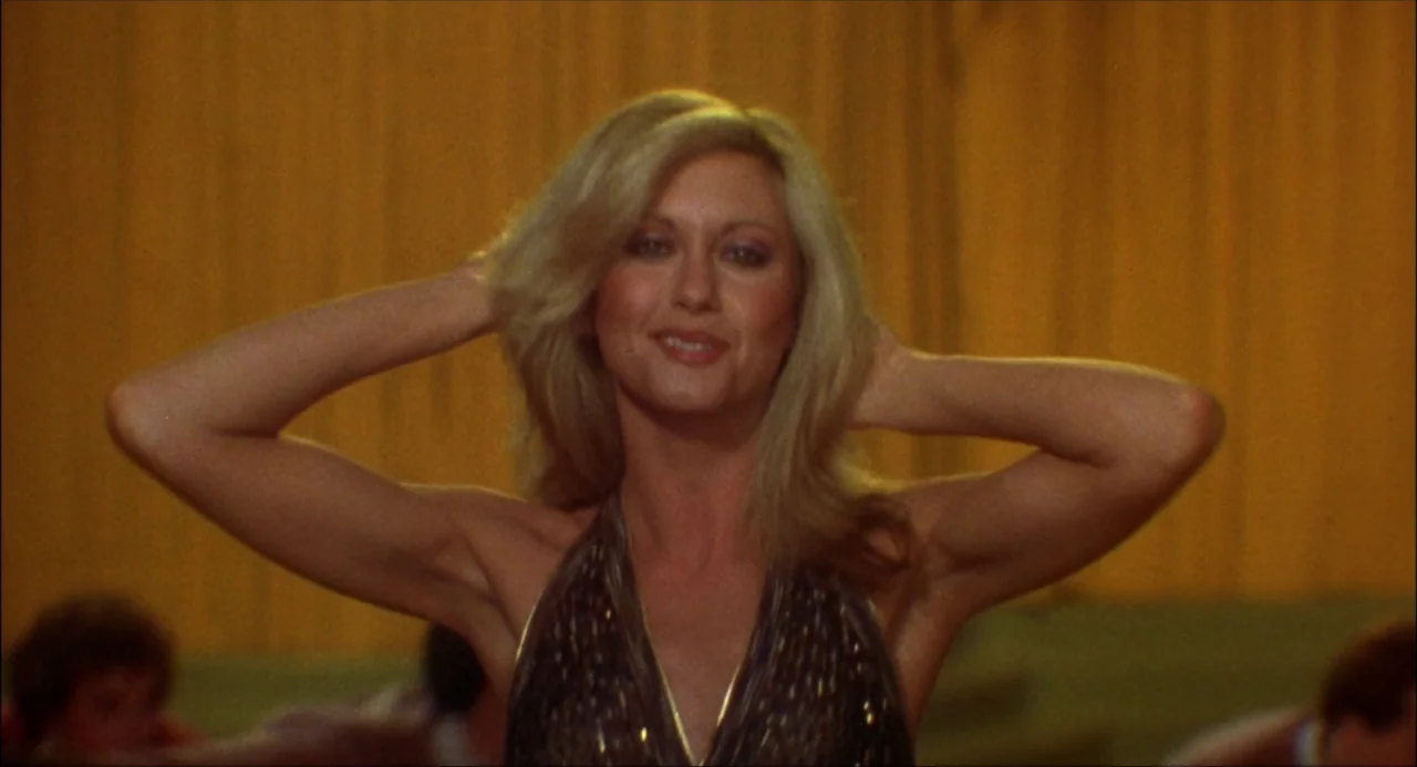 Xanadu (1980) movie still: medium — A woman stands in a bright space with gathered yellow fabric curtains in back. She smiles with both hands placed…