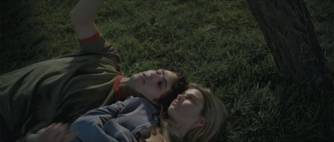 Nahir (2024) movie still: medium — A park setting features green grass beneath a large tree trunk. A young man and a woman lie together on the…
