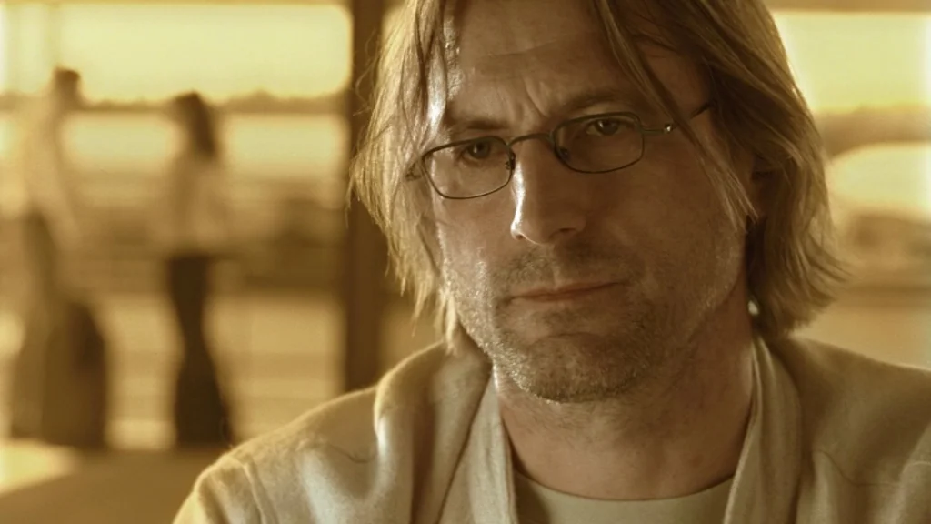 The Ugly Swans (2006) movie still: closeup — A man stands in an indoor area with blurred figures visible in the background. He is centered in the…