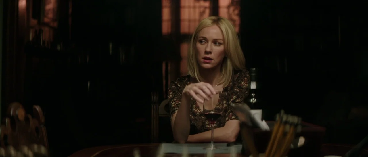The Sea of Trees (2015) movie still: medium — A dark dining room with blurred background silhouettes and wooden furniture. A blonde woman sits at…