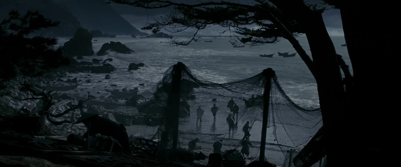 Memoirs of a Geisha (2005) movie still: wide — A rugged, rocky beach at night features dark, crashing waves and small fishing boats. Several…
