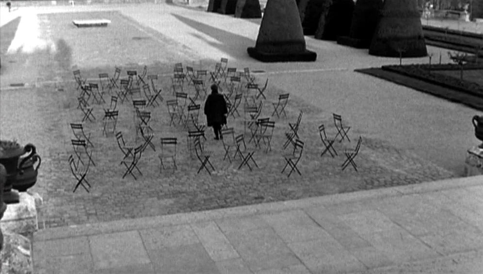 The Tango Lesson (1997) movie still: wide — A formal garden features rows of pruned conical hedges and arranged metal chairs. A man in a long coat…