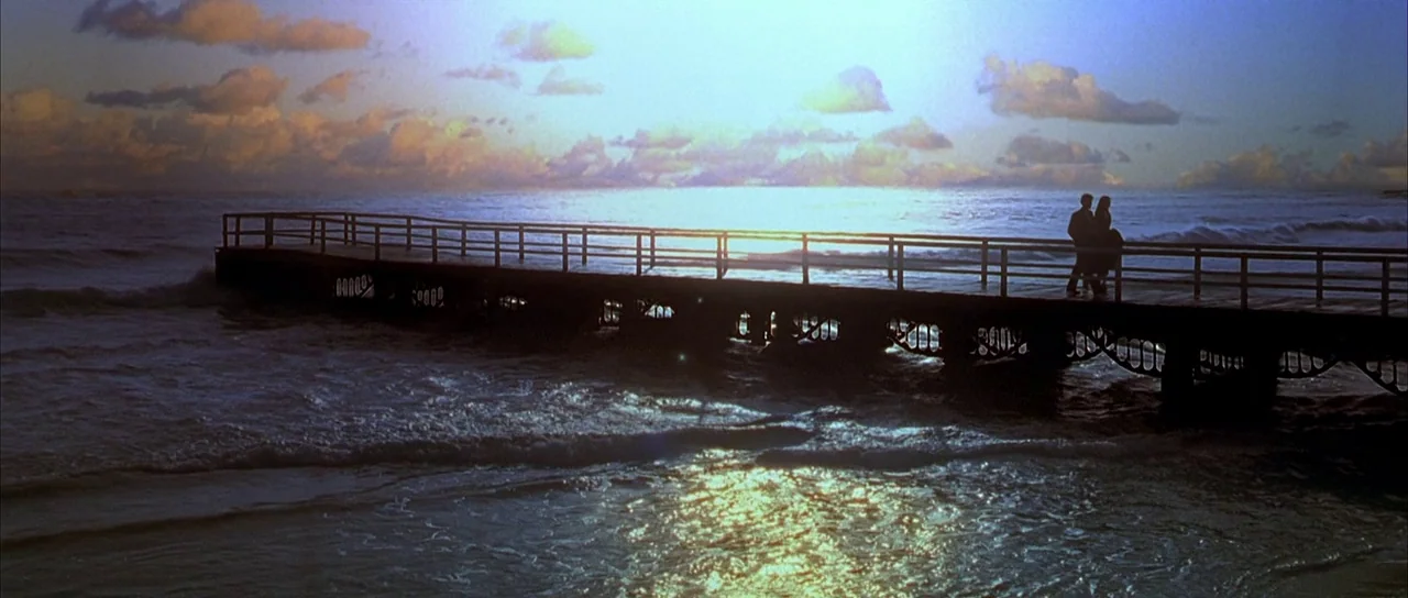 Dark City (1998) movie still: wide — A long wooden pier extends into the ocean under a sunset sky with clouds. A couple stands together at the edge…