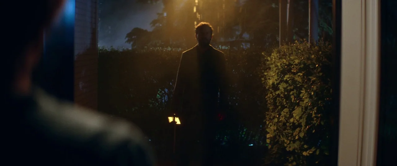 Room For Rent (2017) movie still: medium — A dimly lit outdoor porch area at night with dense hedges behind the subject. A man in a dark jacket…