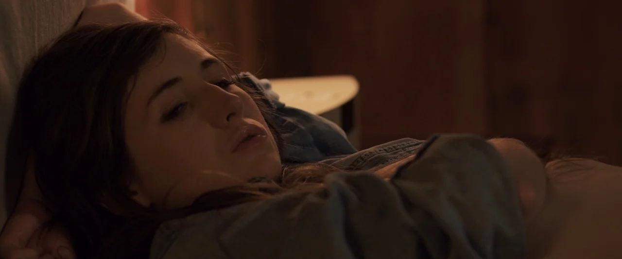 You’re Next (2013) movie still: closeup — A dark indoor space with brown walls and a white side table surface. A young woman reclines in bed while…