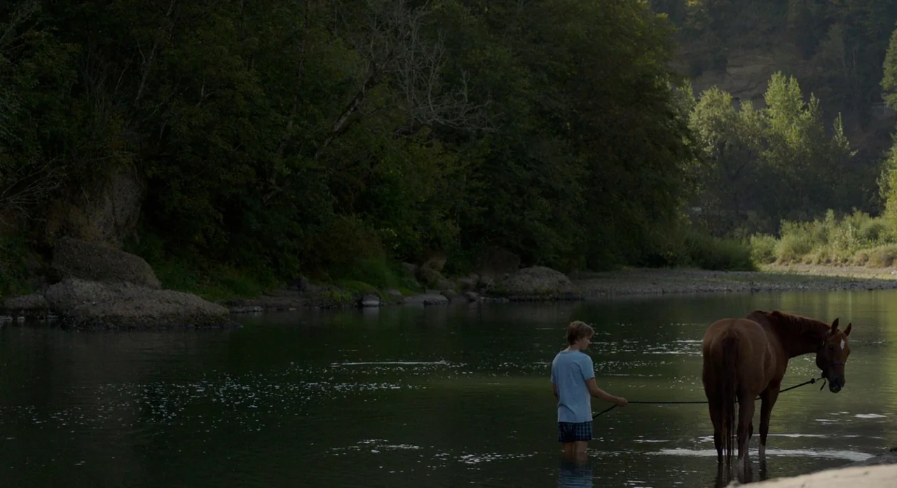 Lean on Pete (2017) movie still: wide — A calm, wide river bordered by dense green trees and rock formations. A young boy in light clothing stands…