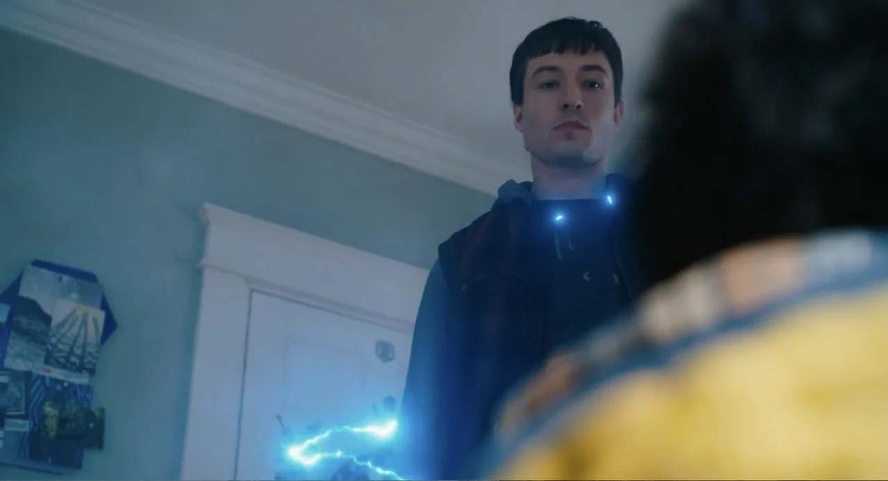 The Flash (2023) movie still: medium — A dimly lit room with light blue walls and a closed white door. A young man stands in front of another person…