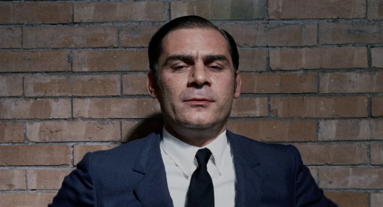 Investigation of a Citizen Above Suspicion (1970) movie still: closeup — A man stands centered directly in front of a weathered brown brick wall. He…