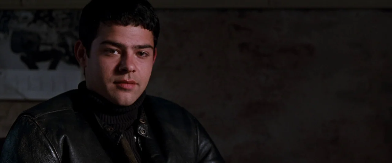 Empire Records (1995) movie still: closeup — A dimly lit interior space with a dark, textured wall behind the subject. A young man wearing a black…