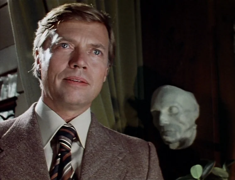 Martha (1974) movie still: medium — A man in a blazer stands before a muted background with heavy drapes. A pale, disembodied plaster head sculpture…