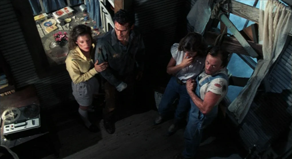 Evil Dead II (1987) movie still: wide — A dimly lit room inside a rustic wooden cabin has dark plank walls. Four people stand together on the floor…