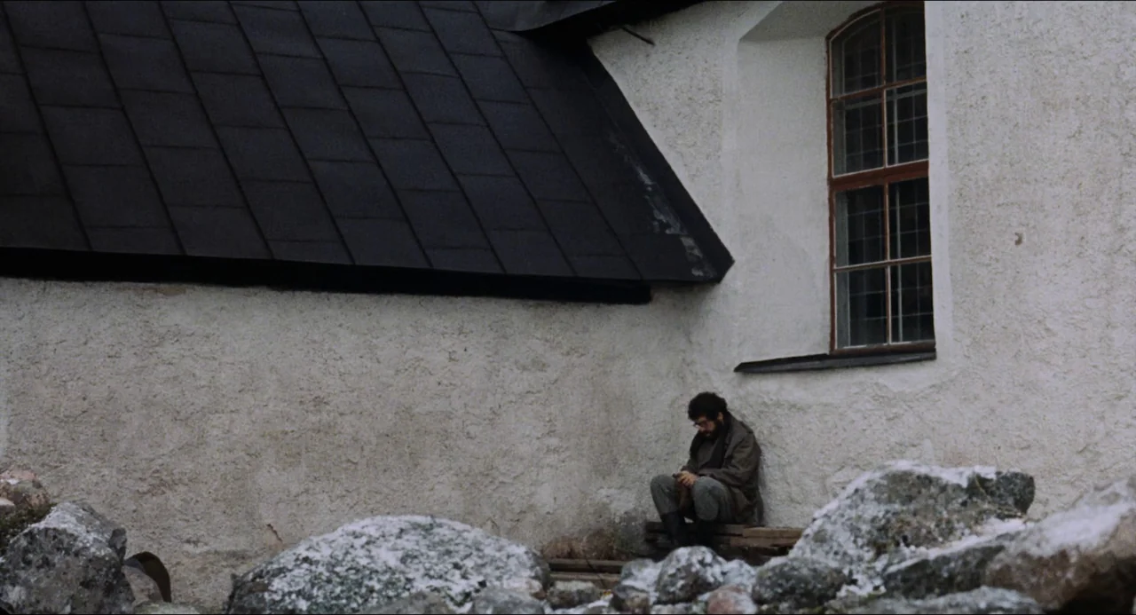 The Touch (1971) movie still: wide — A man sits beside a white stucco wall with a black metal roof corner. The ground is covered in large,…