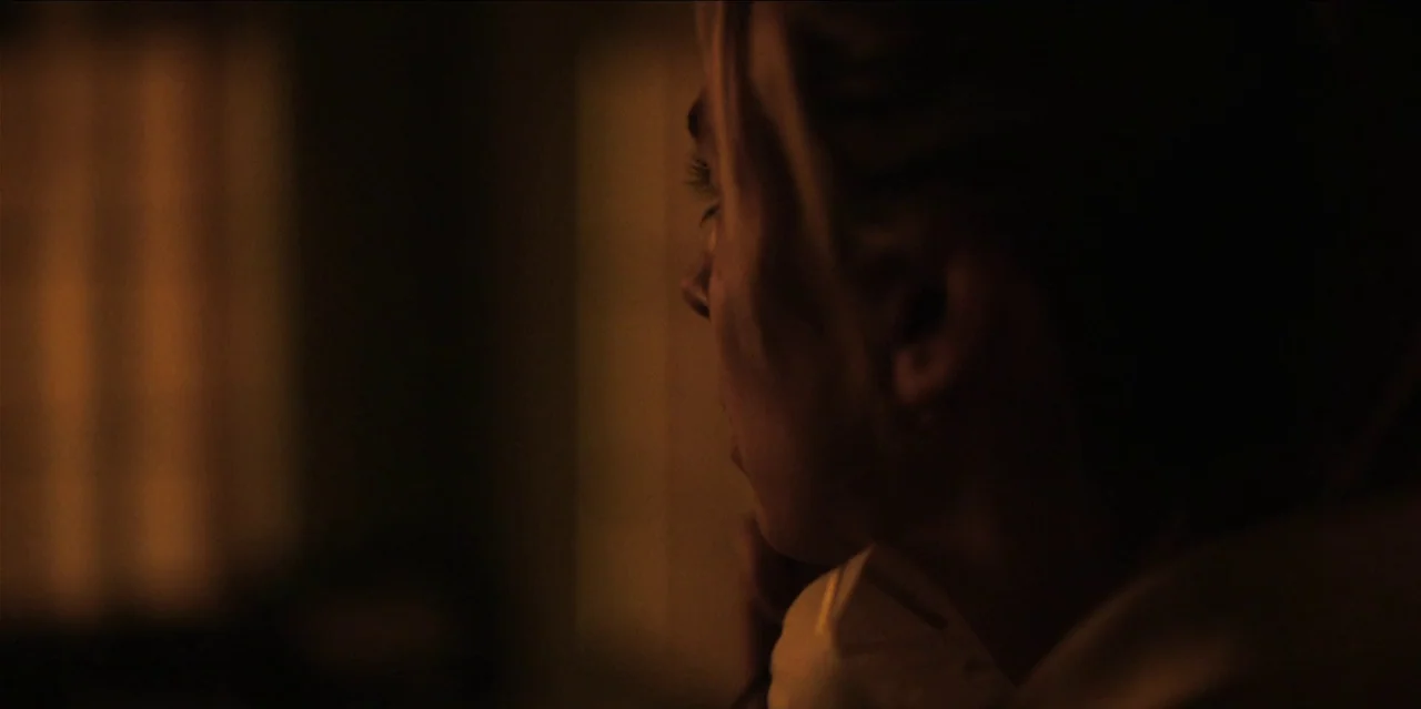 Help (2021) movie still: closeup — A dark indoor space with soft ambient light hitting the subject's profile. A young woman with blonde hair looks…