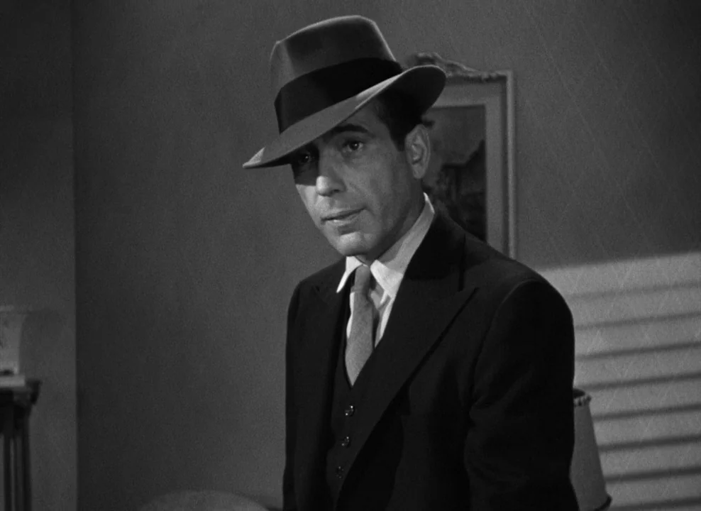 The Maltese Falcon (1941) movie still: medium — An interior space with a dimly lit wall behind a man wearing a hat. The man stands in the…