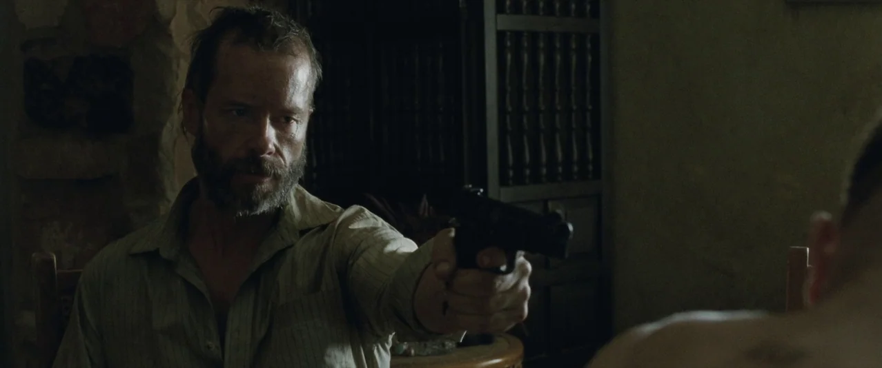 The Rover (2014) movie still: medium — A dimly lit room features a man holding a firearm toward another person. Shadows dominate the background…