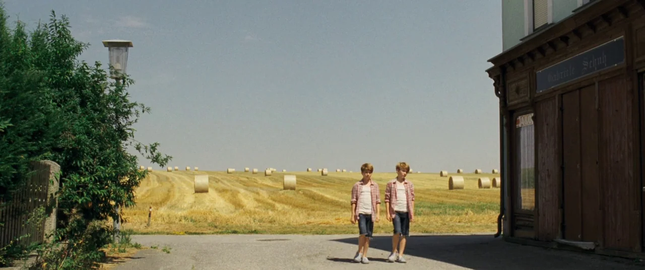 Goodnight Mommy (2014) movie still: fullbody — An asphalt road runs between a leafy bush and a building into a field. Two identical boys walk side…
