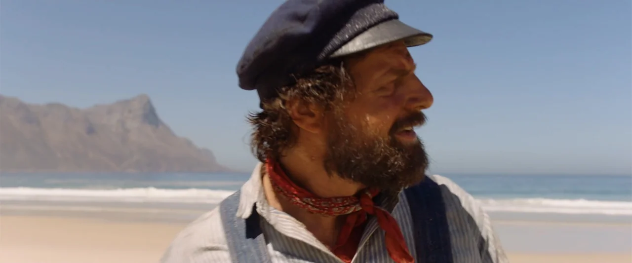 Jim Button and Luke the Engine Driver (2018) movie still: medium — A man stands on a wide, sandy beach under a clear blue sky. The man wears a…