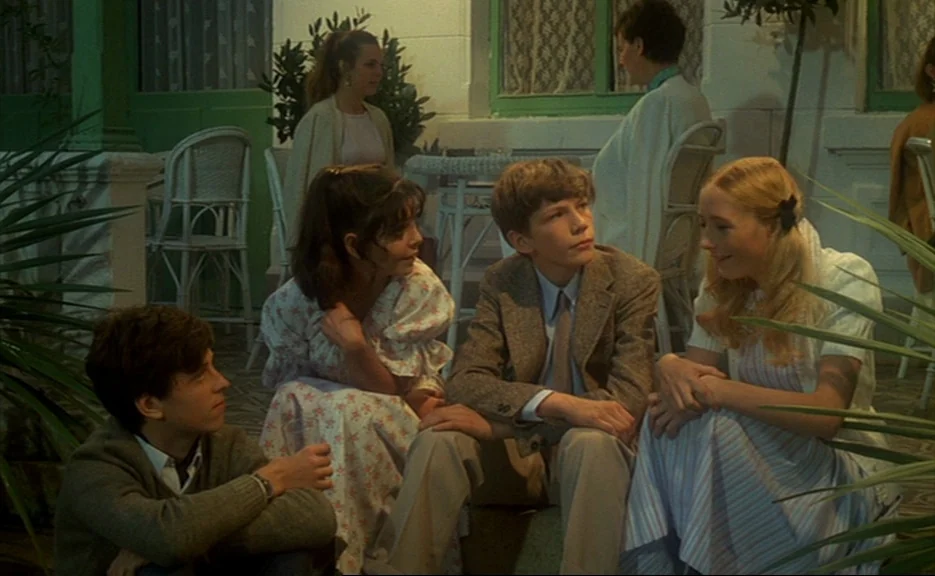Murmur of the Heart (1971) movie still: fullbody — A patio at night with green-painted wood trim and lacy curtains visible behind. Four teenagers…