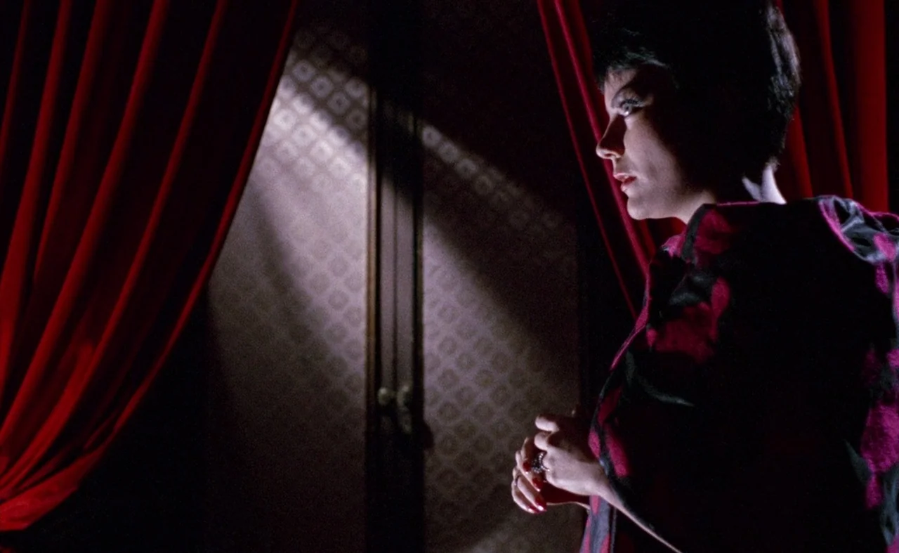Blood And Black Lace (1964) movie still: medium — A dark space features heavy red curtains and a wall with geometric wallpaper patterns. A woman…