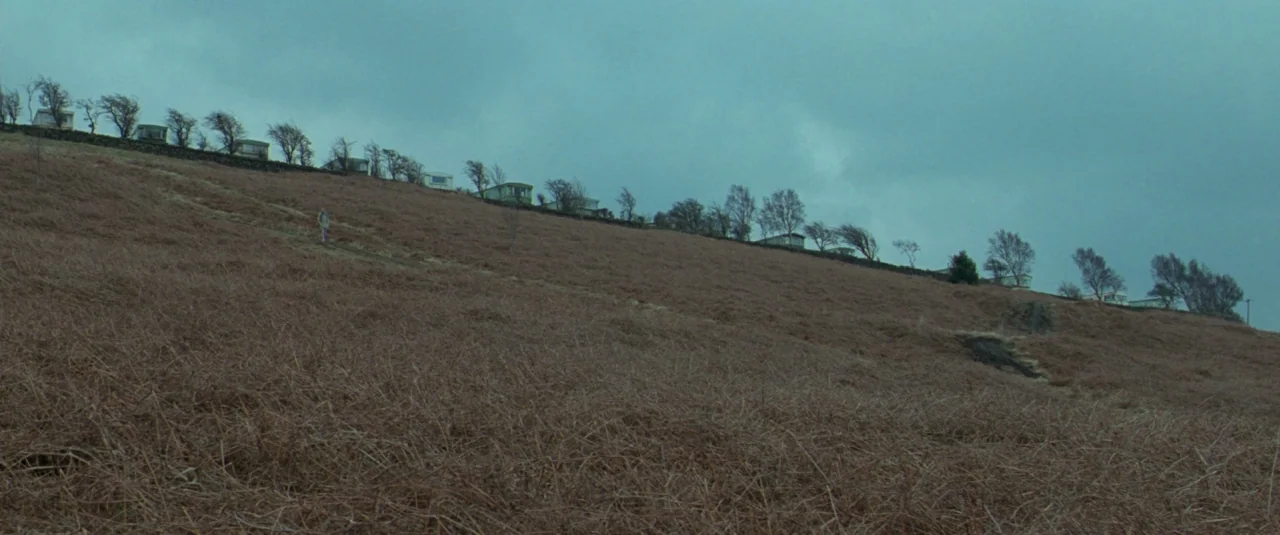 Catch Me Daddy (2014) movie still: wide — A vast, brownish grassy slope rises toward a ridge lined with small mobile homes. Stunted trees and stone…