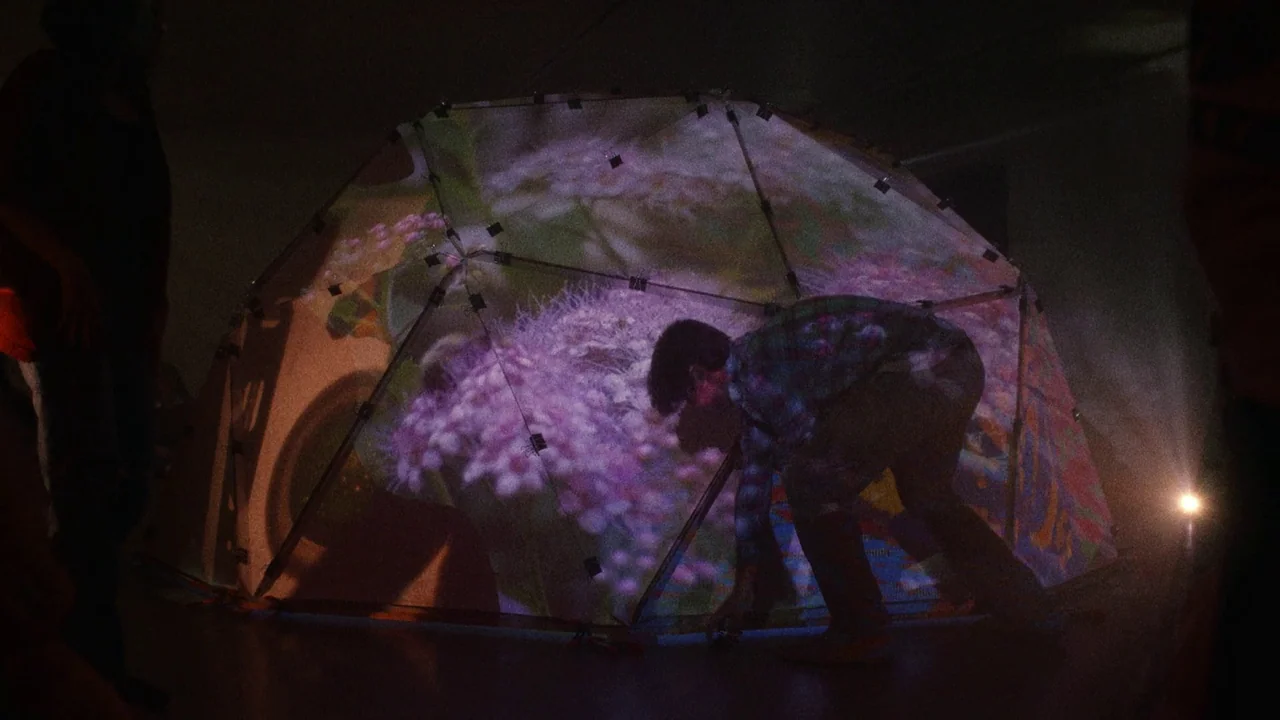 Showing Up (2022) movie still: medium — A dark room contains a translucent geodesic dome structure showing a projected floral image. A man bends…