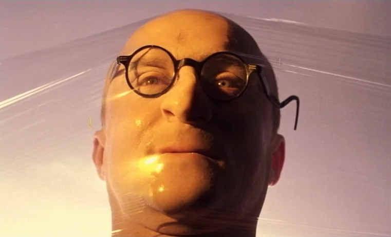 Golem (1980) movie still: closeup — A bald man's face is pressed directly behind a sheet of transparent plastic wrap. He stares ahead while the…