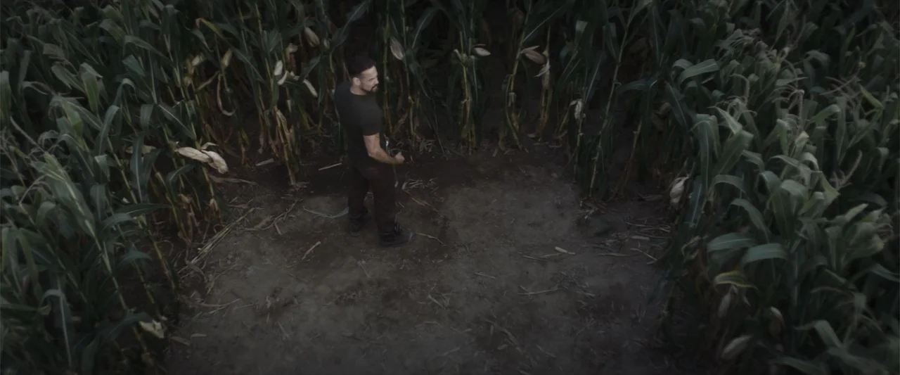 Treblinka's Last Witness (2012) movie still: wide — A clearing surrounded by rows of mature corn stalks under an overcast sky. A man in tactical…