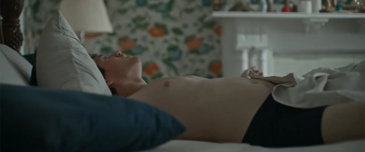 Silent Night, Deadly Night (2025) movie still: medium — A bedroom contains a bed with white sheets and a patterned wallpaper with floral motifs. A…