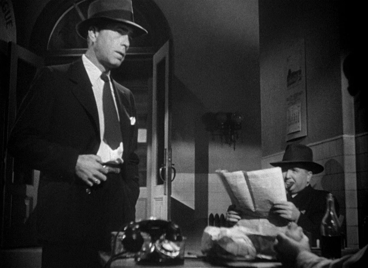 Dead Reckoning (1946) movie still: medium — A dimly lit office features a desk with a rotary telephone and newspapers scattered about. A man in a…