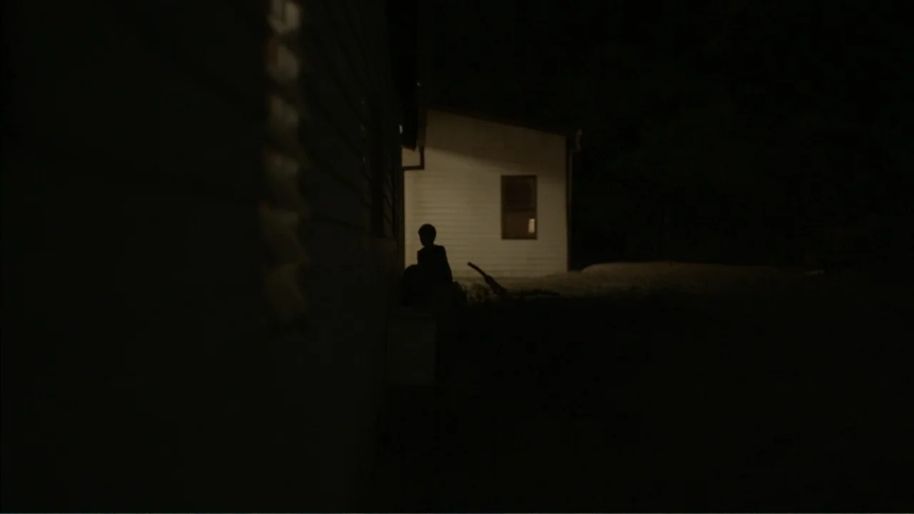 Boy (2010) movie still: wide — A dark yard at night borders a wooden house with one illuminated window. A small child sits on the ground in…