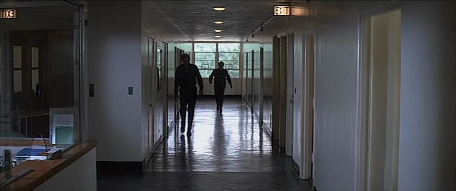 Deliverance (1972) movie still: wide — A long, institutional hallway features white walls, reflective floors, and multiple closed door frames. Two…