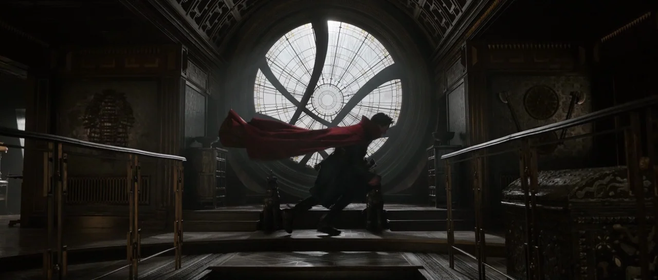 Doctor Strange (2016) movie still: wide — A dark, ornate room with wood-paneled walls features a massive circular window at center. A man in a dark…