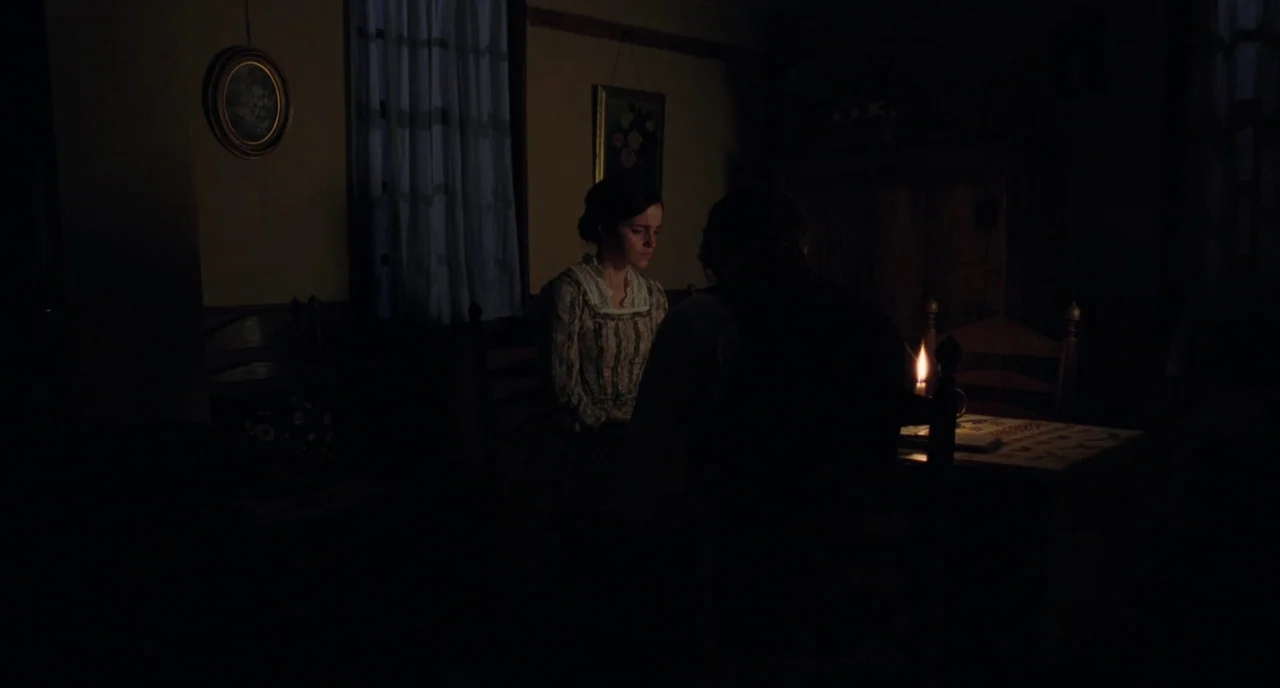 Little Women (2019) movie still: medium — A dimly lit room with simple period furniture and framed wall art. Two people sit at a small table…