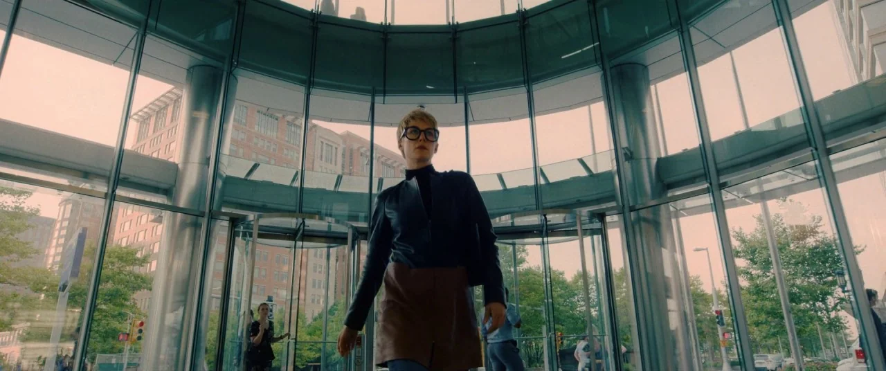 I Care A Lot (2020) movie still: fullbody — A modern building lobby featuring curved glass walls and a central revolving door entrance. A woman with…